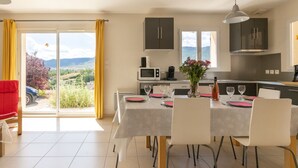 A dining area with a table set for six, a kitchen in the background, and a view of the countryside through a large window. - Les Hauts de Jallia by Interhome (Nyons)