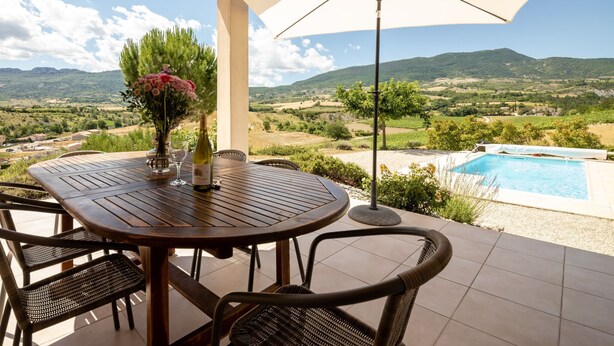 A wooden table with chairs, a bottle of wine, and a vase of flowers on a terrace overlooking a pool and scenic landscape. - Les Hauts de Jallia by Interhome (Nyons)