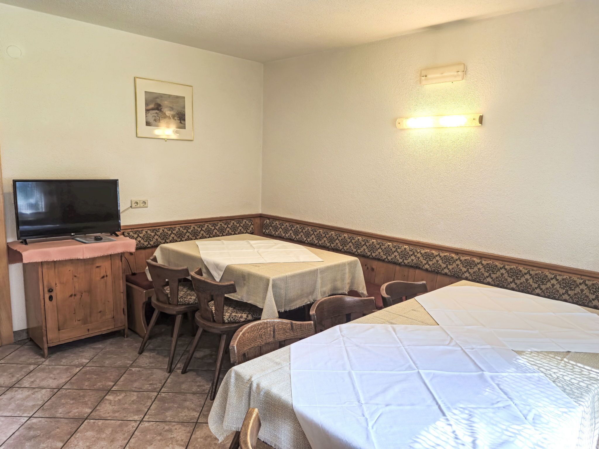 A room with a TV on a wooden cabinet, two tables with white tablecloths, and a patterned wall border.