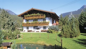 Eine zweistöckige Chalet mit einem geneigten Dach, hölzernen Balkonen und einem kleinen Teich im Vordergrund.