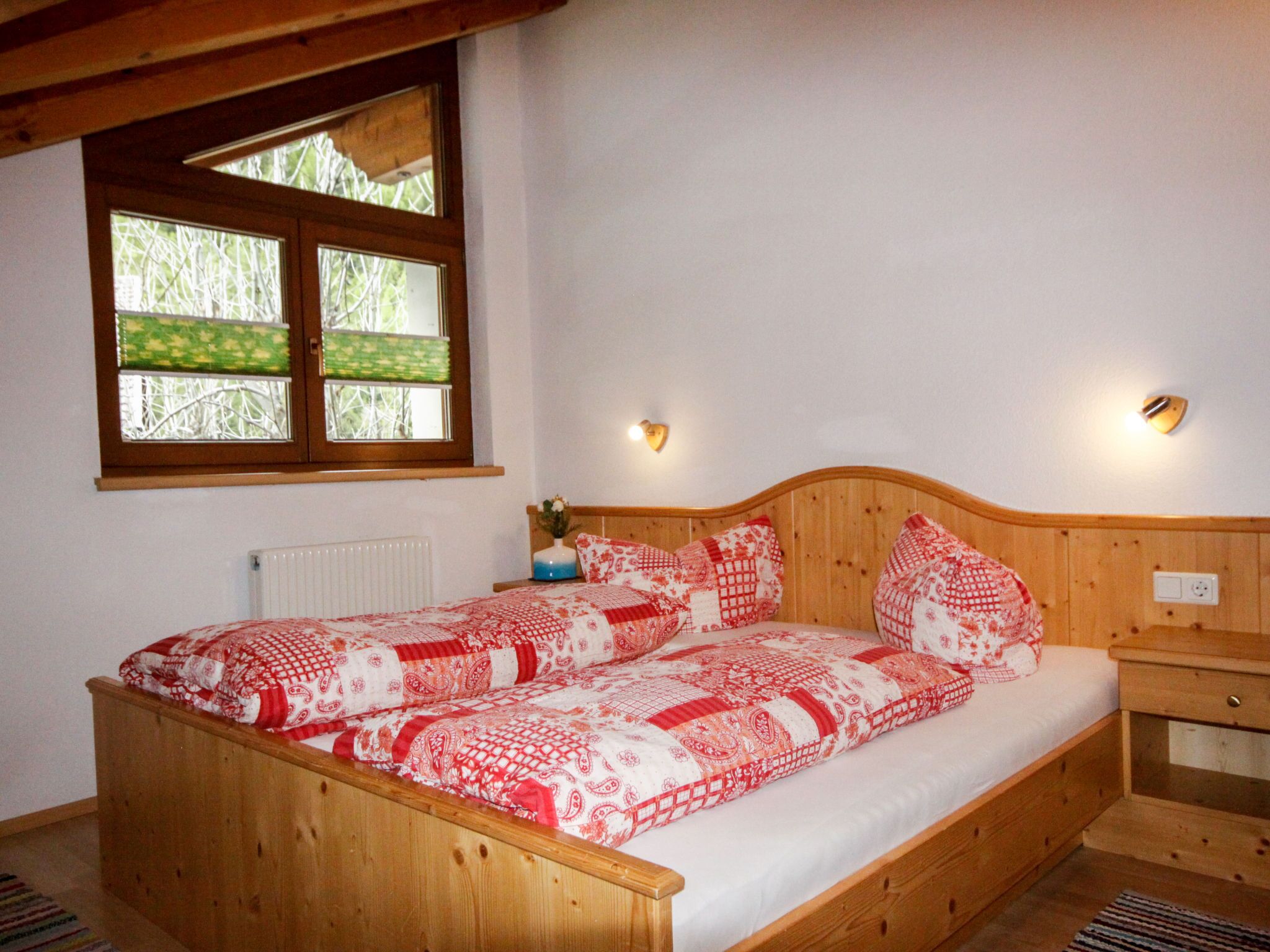 A wooden bed with a red and white patterned bedspread, a nightstand with a vase, and a window with a view of trees.