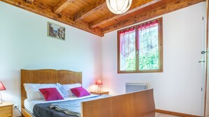 A bedroom with a wooden bed, a nightstand, a lamp, a window with a view of greenery, and a ceiling fan. - Les Galets by Interhome (Valgorge)