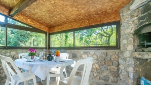 A stone-walled room with a round table and white chairs, a wooden ceiling, and a stone fireplace. - Les Galets by Interhome (Valgorge)