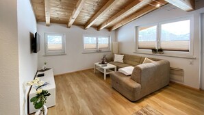 A modern living room with a wooden ceiling, a beige sofa, a white coffee table, and a small side table. There are three windows providing natural light. - Lili - Top 1 by Interhome (Längenfeld)