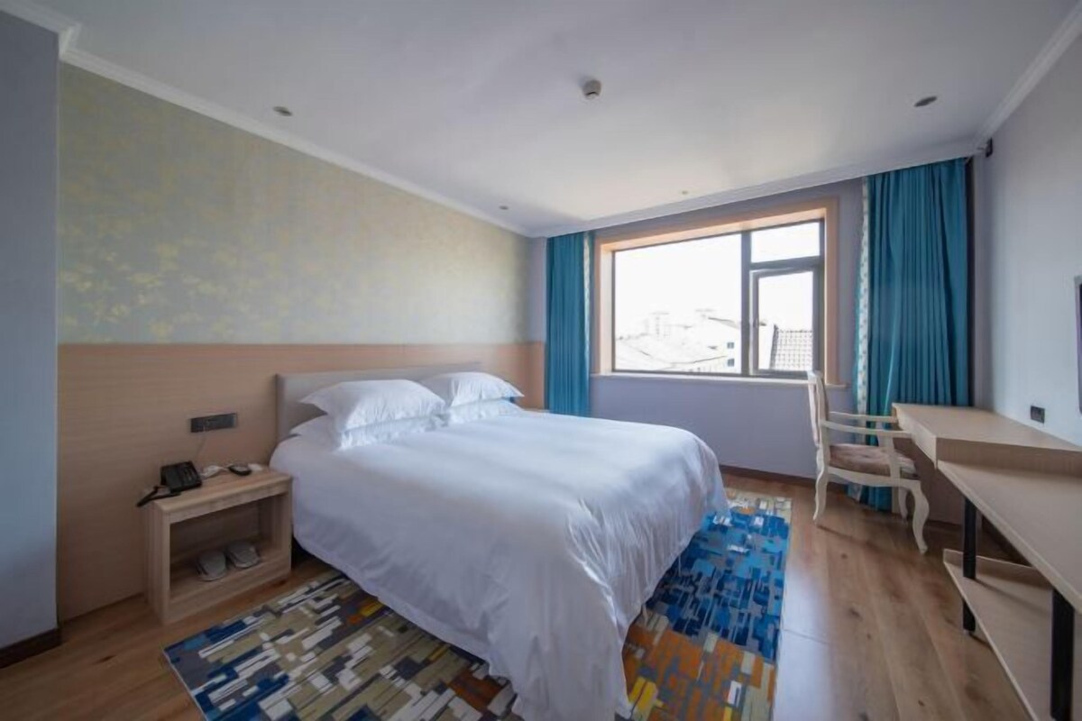 Una habitación de hotel con cama, escritorio, silla, ventana con cortinas, alfombra estampada y mesita de noche con teléfono.