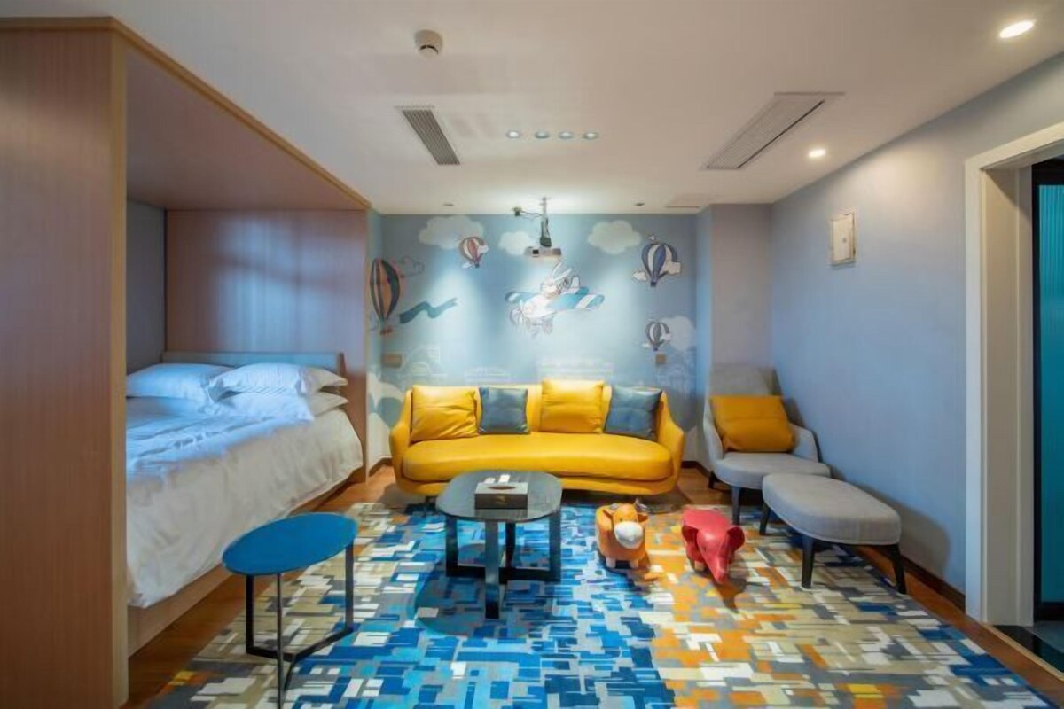 Habitación de hotel pequeña con cama, sofá amarillo, mesita y alfombra con estampado colorido.