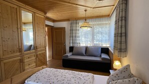 Ein Schlafzimmer mit Holzboden, einem großen Kleiderschrank, einer Schlafcouch, einem Nachttisch mit Lampe und einem Fenster mit Vorhängen.