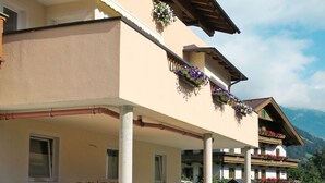 Un bâtiment à deux étages, doté d’un balcon orné de fleurs, d’un mur de pierre à l’avant et, en arrière-plan, d’un ciel dégagé.