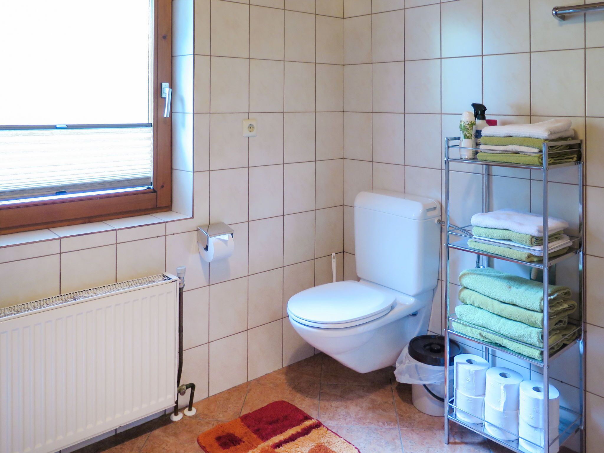 A small bathroom with a toilet, a radiator, a shelf with towels, and a window.