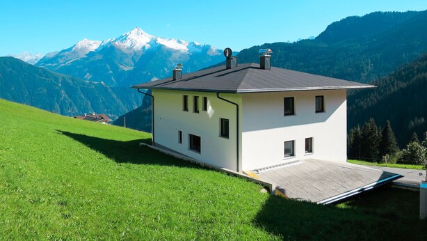 Ein eingeschossiges Haus mit geneigtem Dach, gelegen auf einem grasbewachsen Hügel mit Blick auf einen Berg.