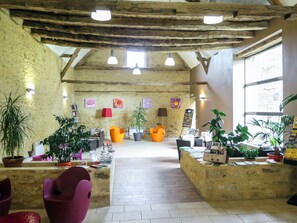 Ein rustikaler Innenraum mit freiliegenden Holzbalken, einer zentralen Sitzgruppe mit bunten Stühlen und verschiedenen topfpflanzengefüllten Blumen.