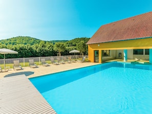 A swimming pool with lounge chairs and umbrellas, surrounded by a wooden deck and a building with a red roof.