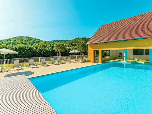 A swimming pool with lounge chairs and umbrellas, surrounded by a wooden deck and a building with a red roof.