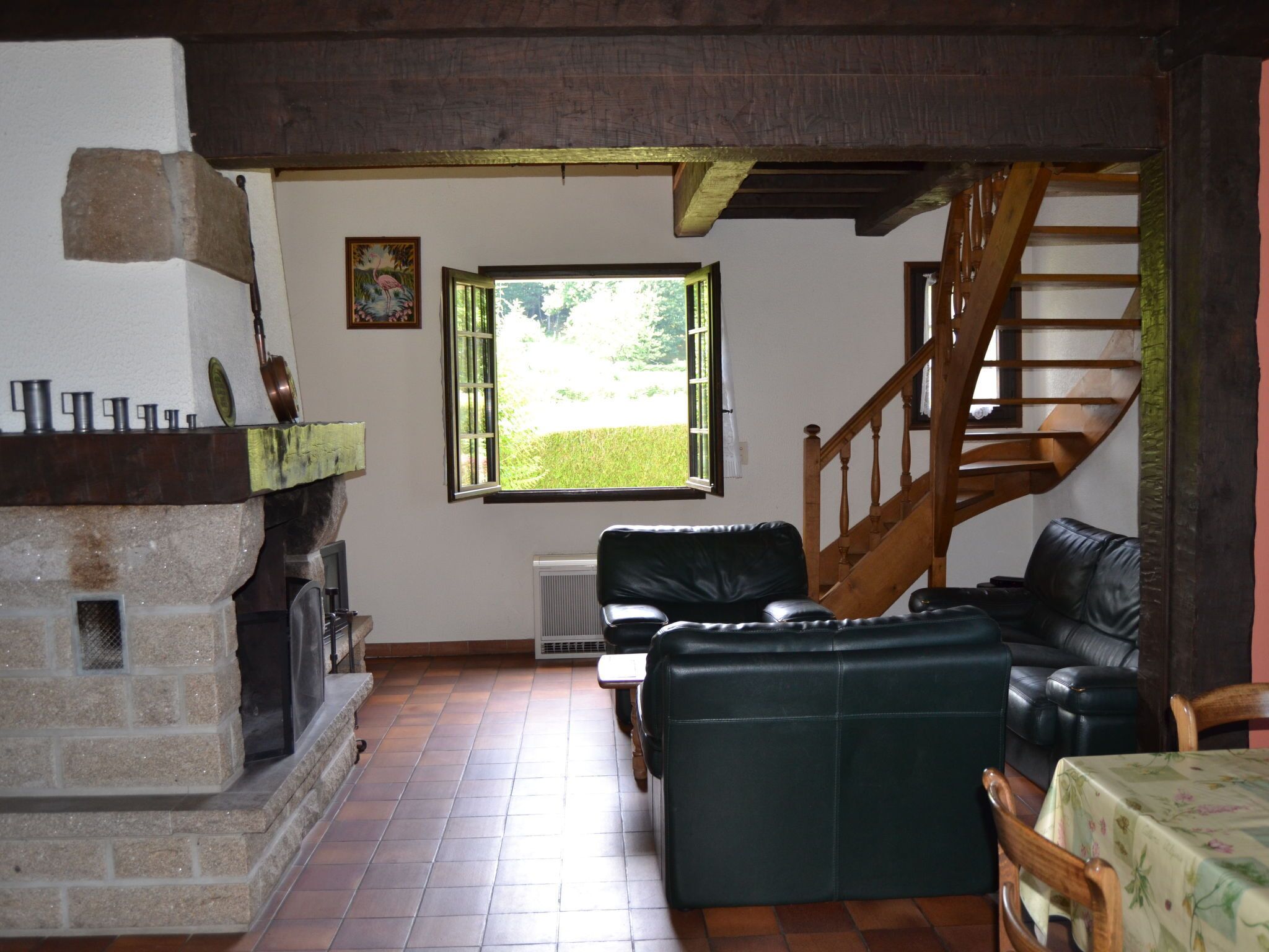 A cozy living room with a fireplace, a green sofa, a wooden staircase, and a window with a view of greenery.