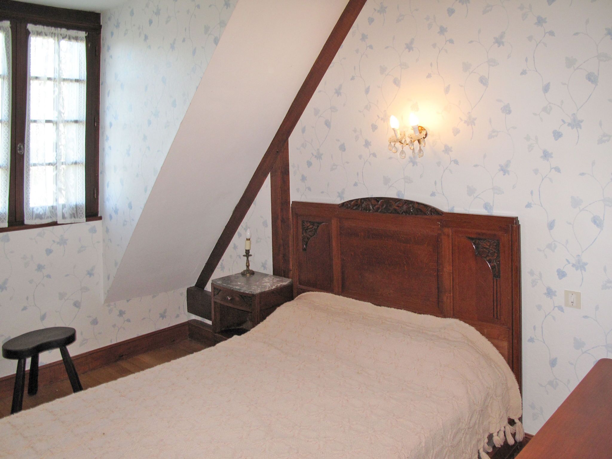 A bedroom with a sloped ceiling, a wooden bed frame, a nightstand with a lamp, and floral wallpaper.