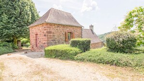 A stone building with a sloped roof, surrounded by greenery and a gravel path.