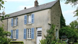 A two-story house with blue shutters, a white door, and a garden with white metal chairs and a table.