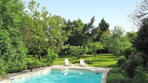 A rectangular swimming pool with a clear blue water surface, surrounded by a concrete edge. There are two white lounge chairs positioned on the pool's edge, facing the water. The pool is set in a lush, green landscape with various trees and shrubs.