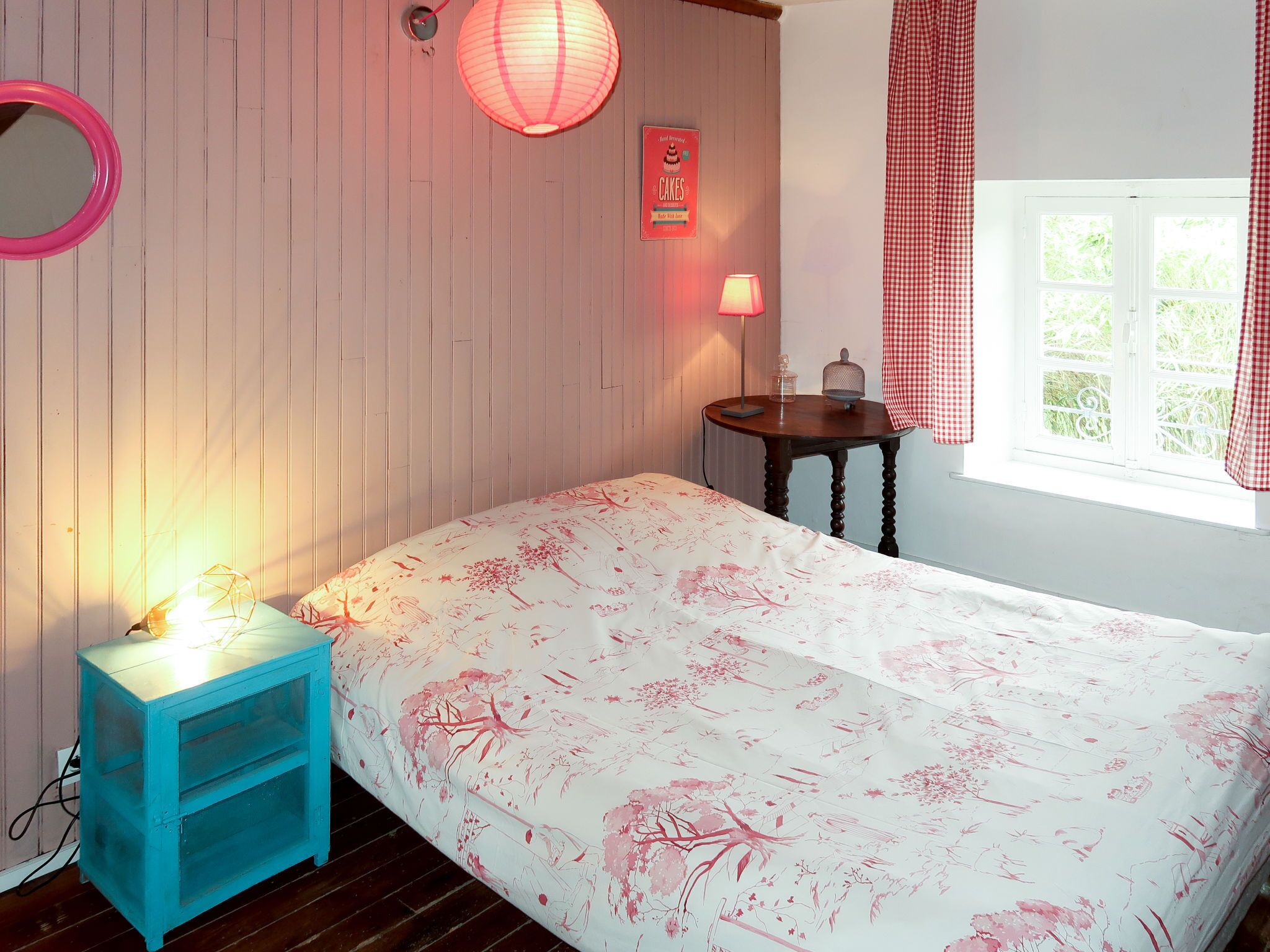 A bedroom with a bed, a nightstand, a lamp, a wall clock, a window with curtains, and a wall-mounted sign.