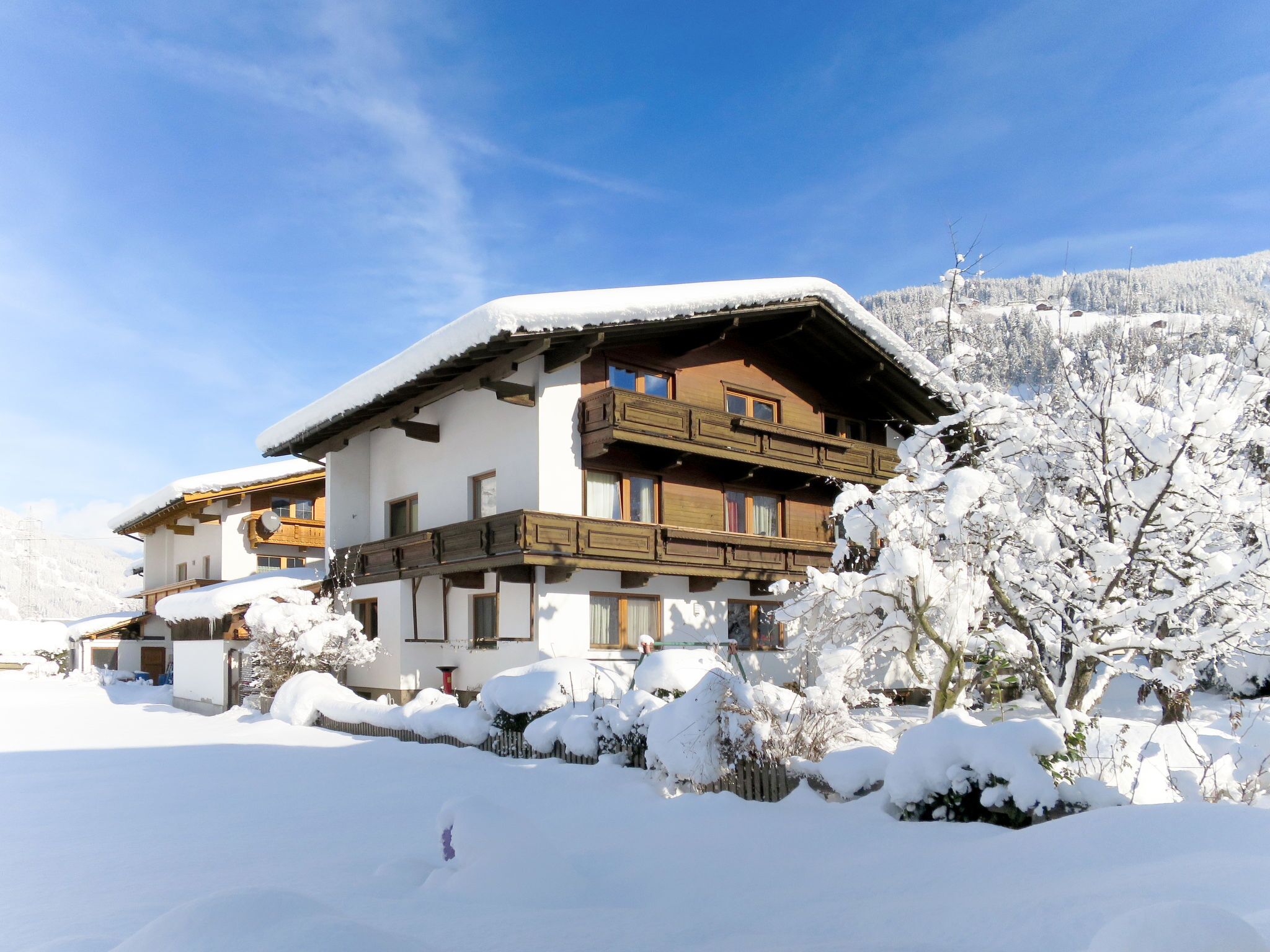 A two-story chalet with a balcony, surrounded by snow-covered trees and a mountain in the background.