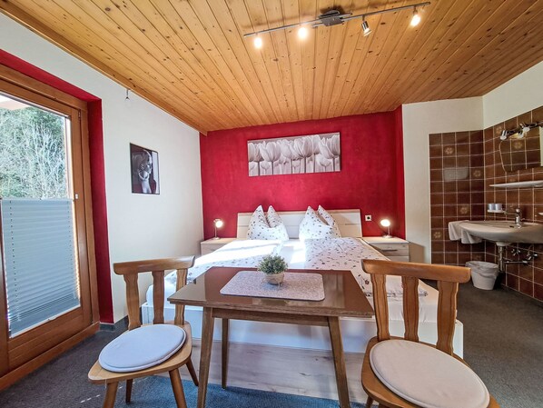 A bedroom with a wooden ceiling, a red accent wall, a bed with white bedding, a bedside table with a lamp, and a bathroom with a sink and tiled walls.