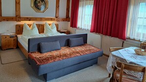 Een slaapkamer met een bed, een slaapbank, een houten hoofdbord, een nachtkastje, een tafel met een mand, en een raam met rode gordijnen.