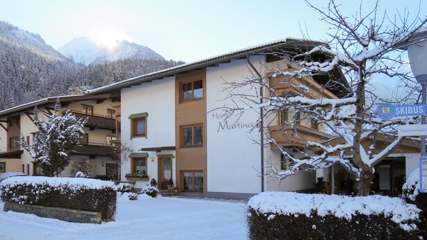 Un hotel con ingresso innevato, un cartello con la scritta 'Skibus' e montagne sullo sfondo.