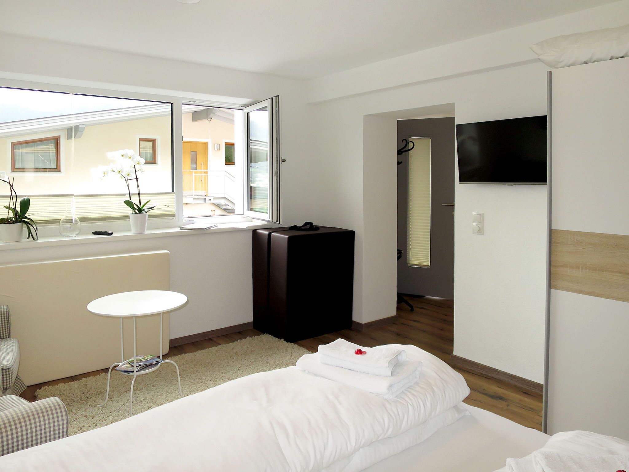 Una habitación de hotel con una cama, una pequeña mesa redonda, una ventana con plantas, una maleta y un televisor en la pared.