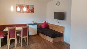 Une pièce compacte comprenant une table à manger en bois, des chaises rouges, un petit coin cuisine, un canapé-lit, un téléviseur fixé au mur et une horloge murale.