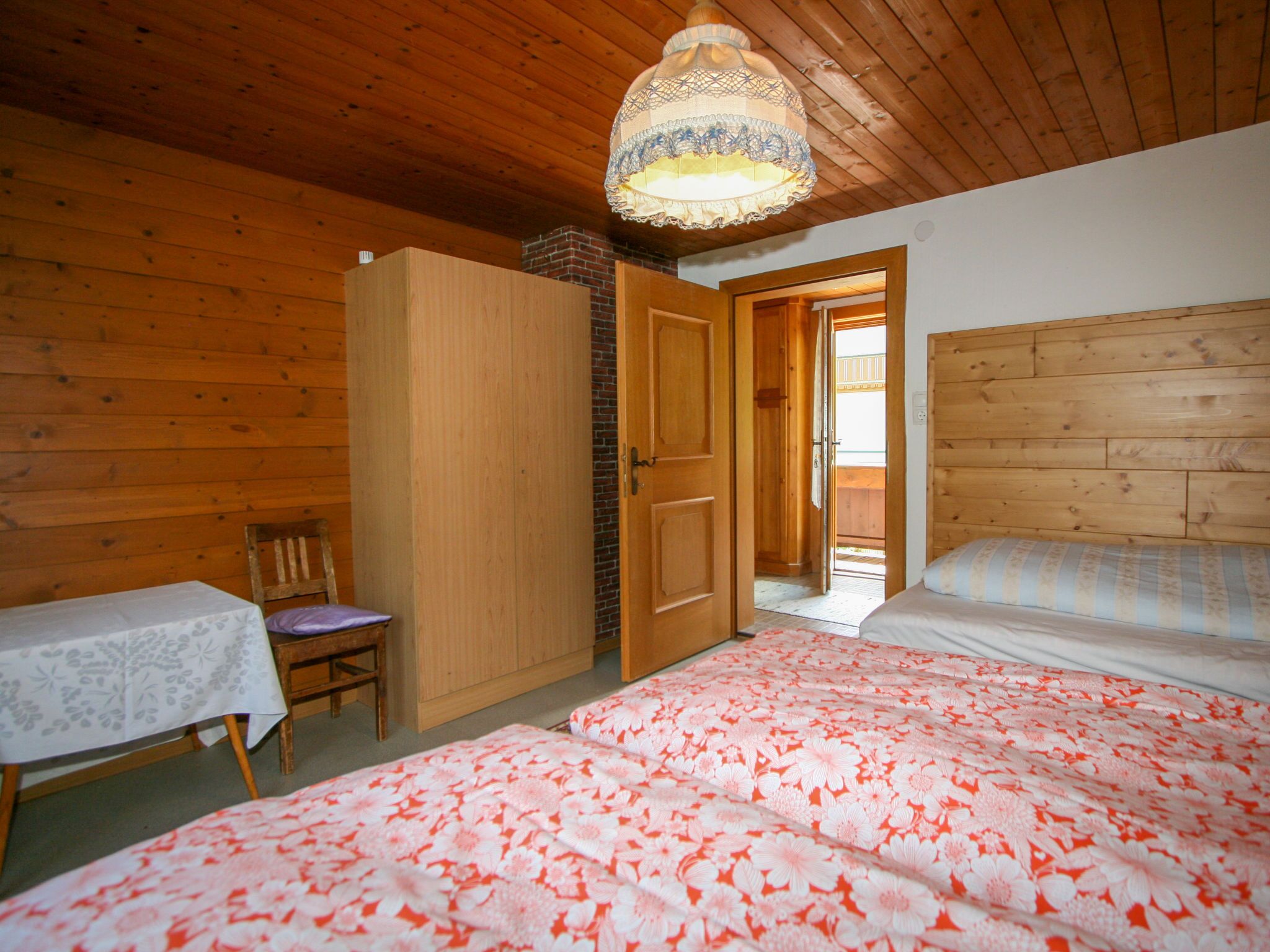 Ett sovrum med träväggar, en säng med blommigt sänglinne, en trägarderob, ett litet bord med en duk och en stol.