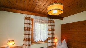 A cozy room with wooden ceiling beams, a bed with white bedding, two bedside tables with lamps, and a window with patterned curtains.