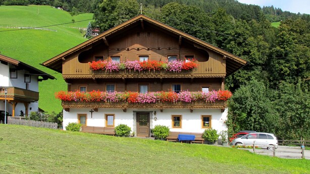A two-story wooden house with balconies adorned with colorful flowers, set against a backdrop of green hills and a clear sky.