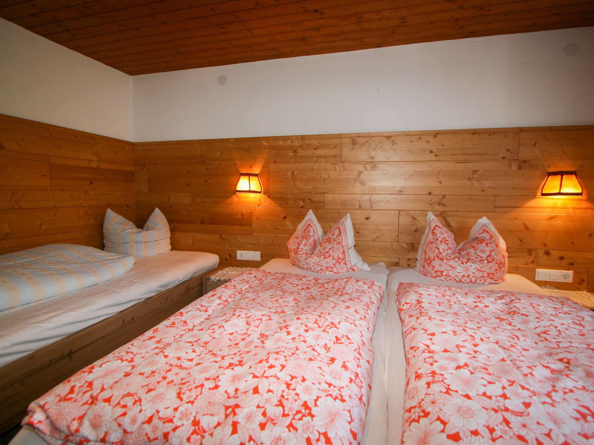 Ein Zimmer mit zwei Betten, jeweils mit floralem Bettbezug, Holzwänden und Wandleuchten.