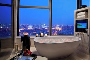 A modern bathroom with a large bathtub filled with rose petals, a city view, and a vanity with a television. - Tangla Hotel Tianjin (TIANJIN)