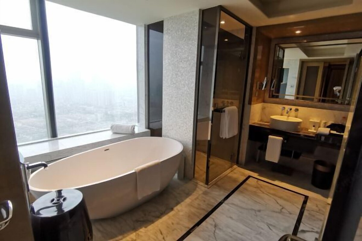 A modern bathroom with a freestanding bathtub, a glass-enclosed shower, a large mirror, and a view of the city.