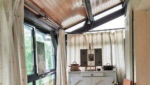 A cozy room with a wooden ceiling, a large window, a white cabinet with kitchen items, and two chairs with cushions.