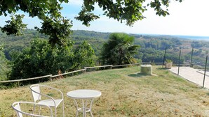 An outdoor seating area with two white metal chairs and a table, surrounded by a grassy field with scattered leaves, overlooking a hilly landscape with trees and a distant view of a body of water. - Roque Piquet by Interhome (L'Honor-de-Cos)