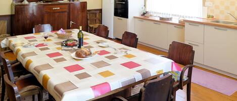 Un comedor con una mesa dispuesta para cuatro personas, rodeada de sillas de madera. Se ve una cocina con armarios blancos y un refrigerador al fondo.