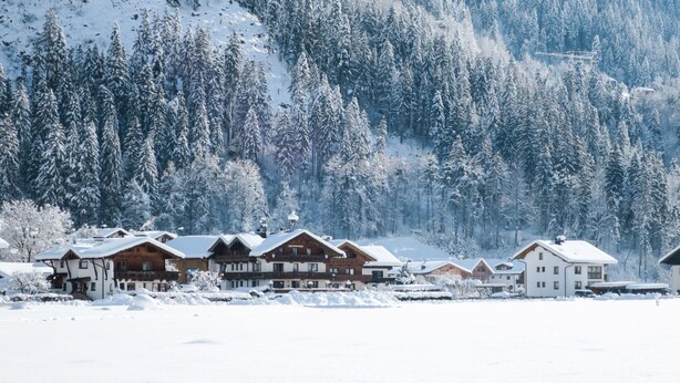 A row of wooden cabins with snow-covered roofs, set against a backdrop of a snow-covered forest and mountains.