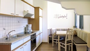 A modern kitchen with white cabinets, a built-in oven, and a coffee maker. There is a dining area with a bench and chairs.