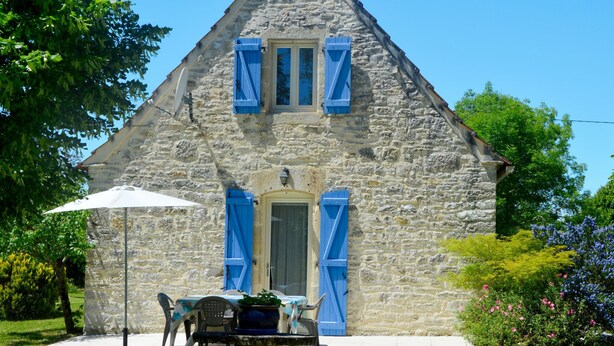 Un edificio de piedra con techo triangular, contraventanas azules y una puerta. Hay una mesa y sillas afuera con una sombrilla blanca.