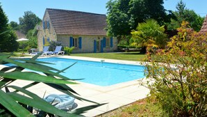 Una piscina con sillones de descanso, un edificio de piedra con contraventanas azules y abundante vegetación.