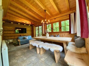 A wooden cabin interior with a stone floor, a wooden dining table, benches, a sofa, and a TV mounted on the wall.