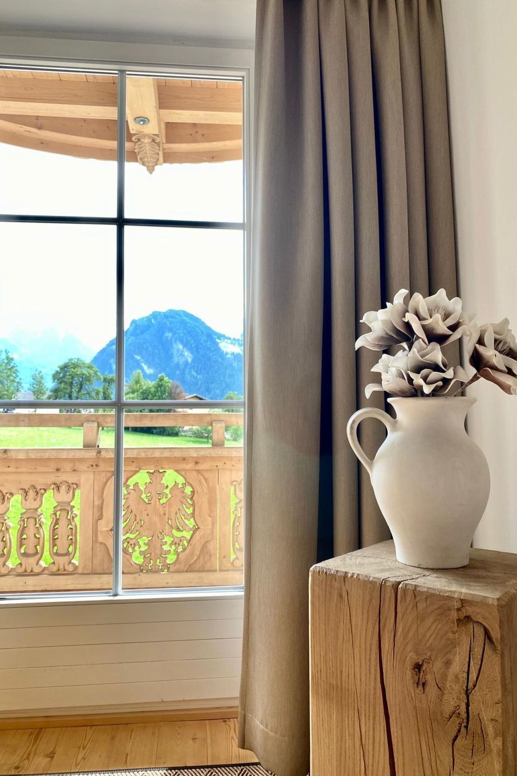 A room with a large window, a wooden floor, a white vase with flowers, and a curtain.