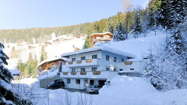 Un hotel de varios pisos con tejado cubierto de nieve, rodeado de árboles y otros edificios en una zona montañosa.