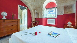 Un dormitorio con techo de piedra, paredes rojas, cómoda de madera, cama con colcha blanca, ventana con vistas y una pequeña mesa con lámpara.