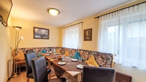 Un coin repas avec une longue table, des chaises et un banc. Deux tableaux ornent les murs, ainsi qu’un lampadaire et une fenêtre avec des rideaux.