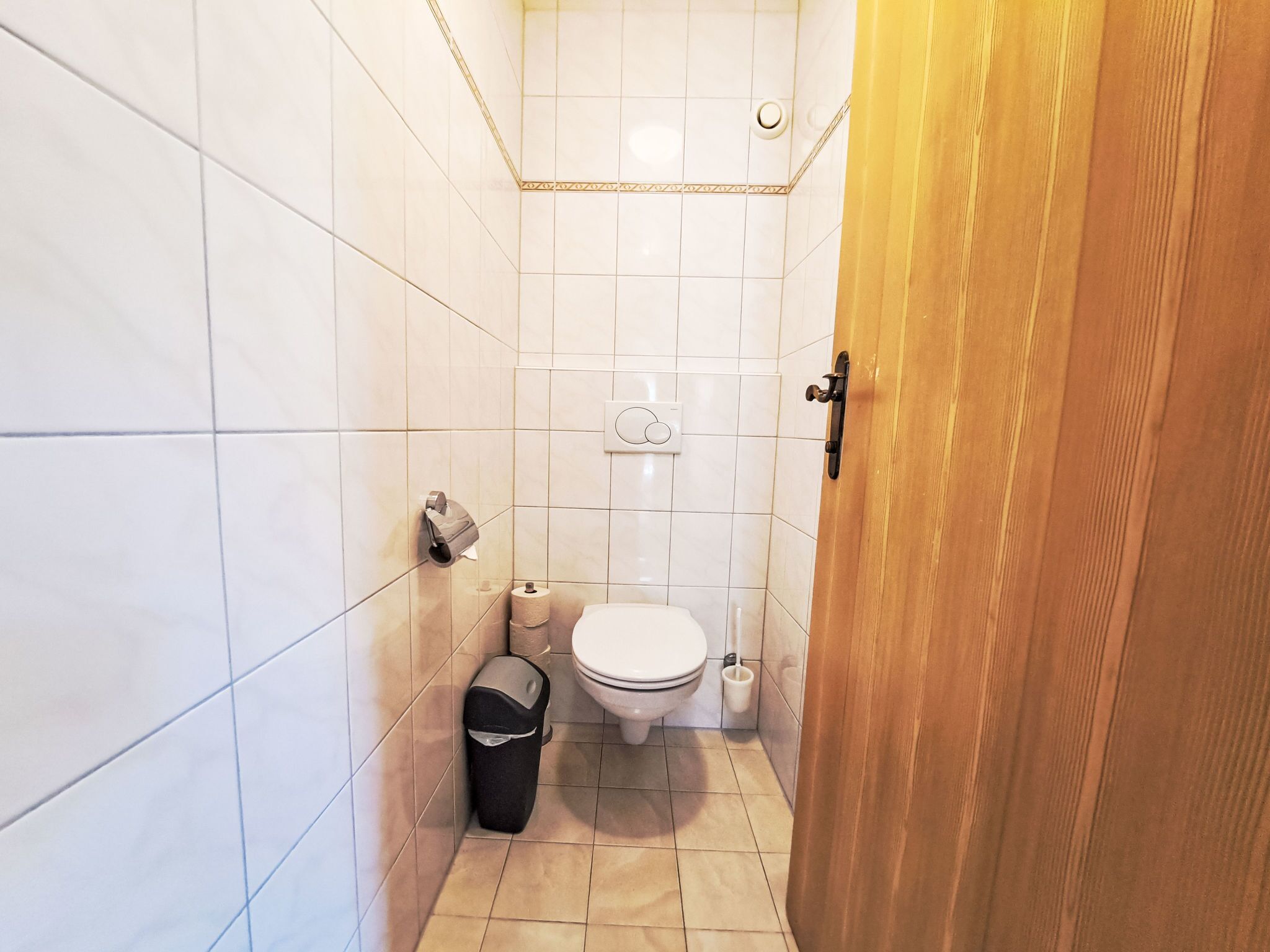 A small bathroom with white tiles, a toilet, a trash bin, and a toilet paper holder.