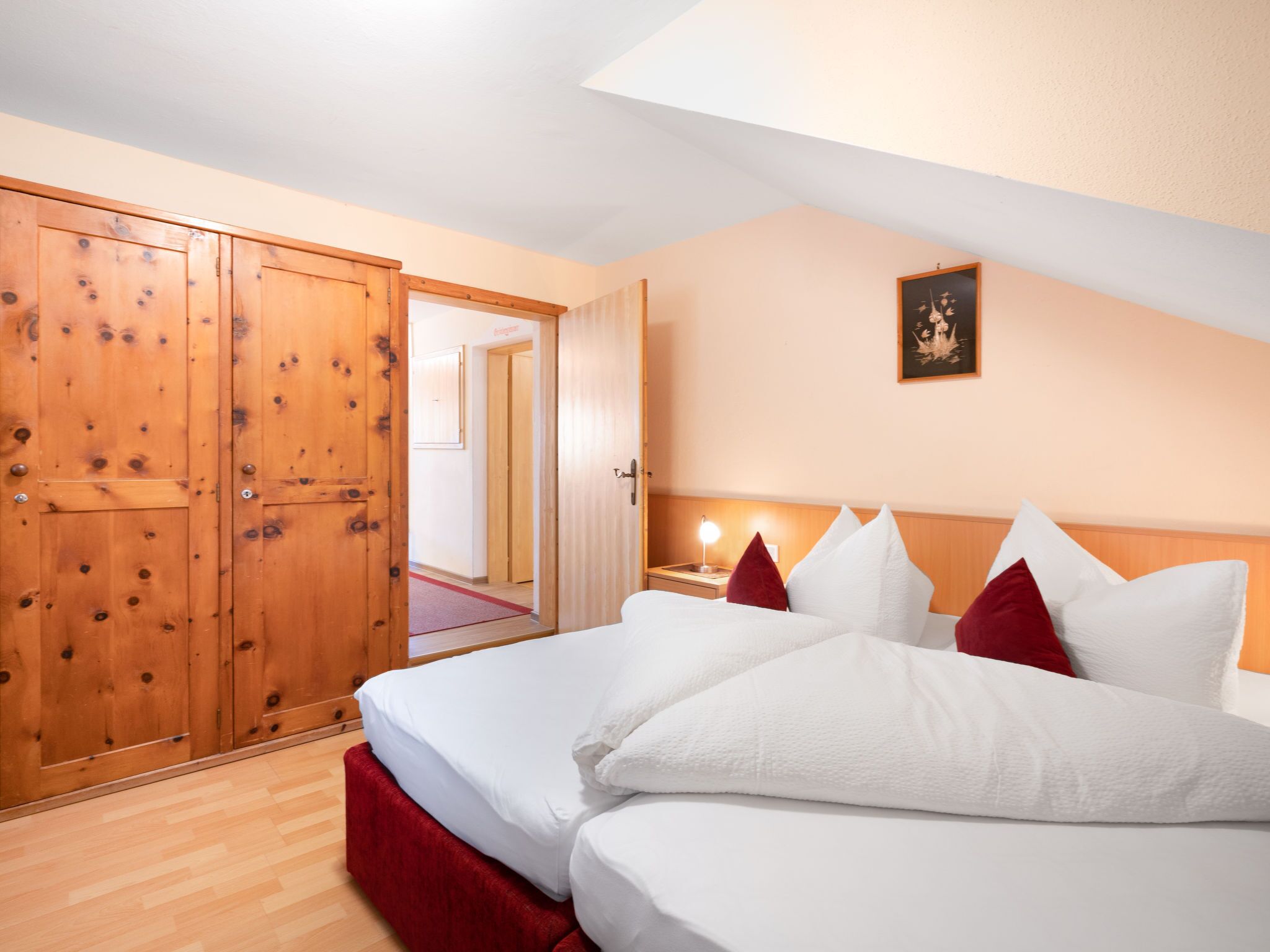 A hotel room with a wooden wardrobe, a bed with white bedding and red pillows, a nightstand with a lamp, and a framed picture on the wall.