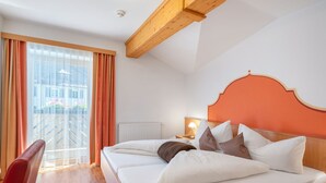 A hotel room with a bed, a nightstand, a lamp, a window with curtains, and a view of buildings outside. - Apartment Rosa by Interhome (Mayrhofen)